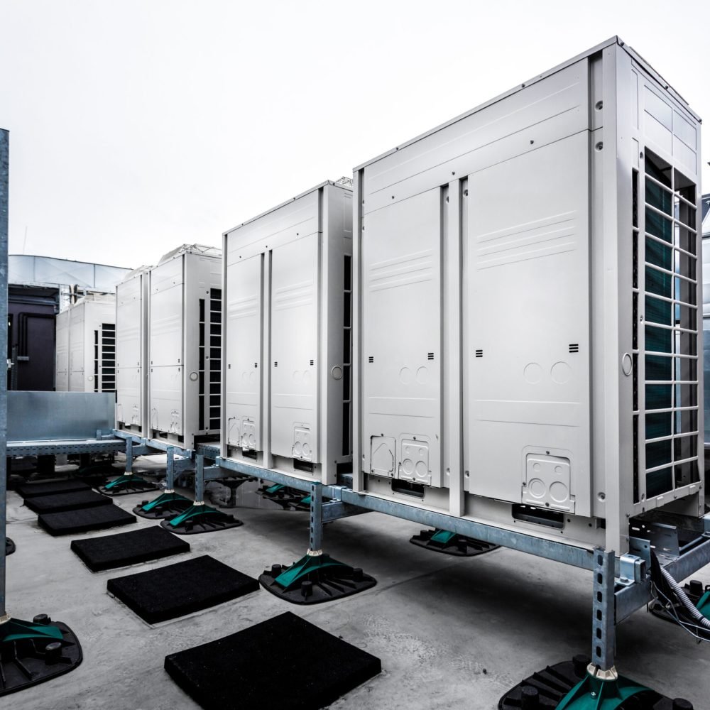 Series of gradually receding air conditioning units on roof with other parts of ventilation system. Sky is cloudy in uniform gray color. Focus is on the first unit with slight fall off in background.
